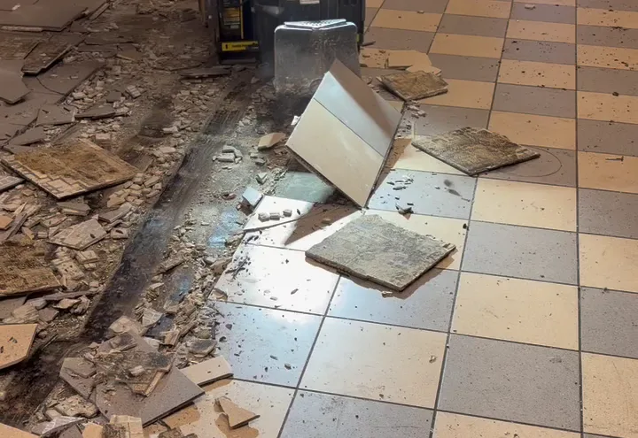 Ride-on scraper blade flipping a ceramic floor tile loose, broken tile piled beside it