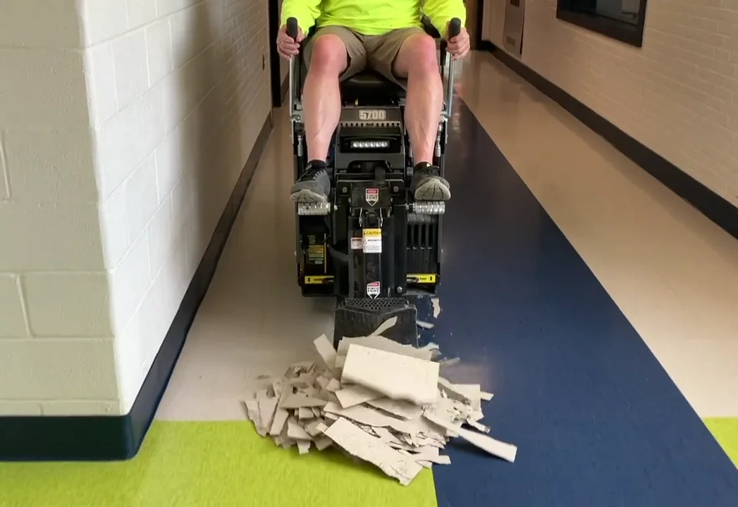 Ride-on floor scraper popping VCT tile in a long hallway, the removed tiles piled ahead of the blade